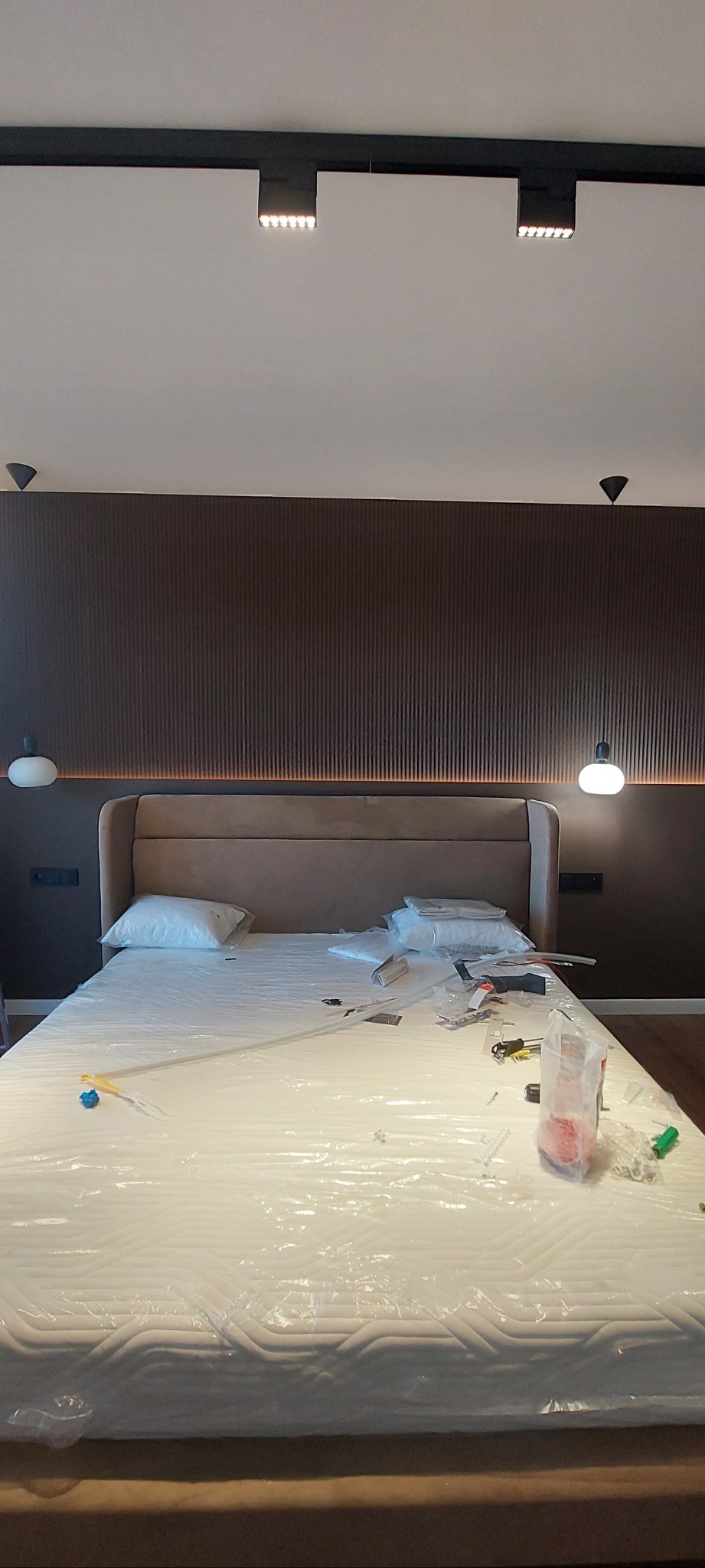 Montaż łóżka z beżowym zagłówkiem na tle ściany z dekoracyjnymi panelami. Widoczne narzędzia i folia ochronna na materacu. Oświetlenie sufitowe.