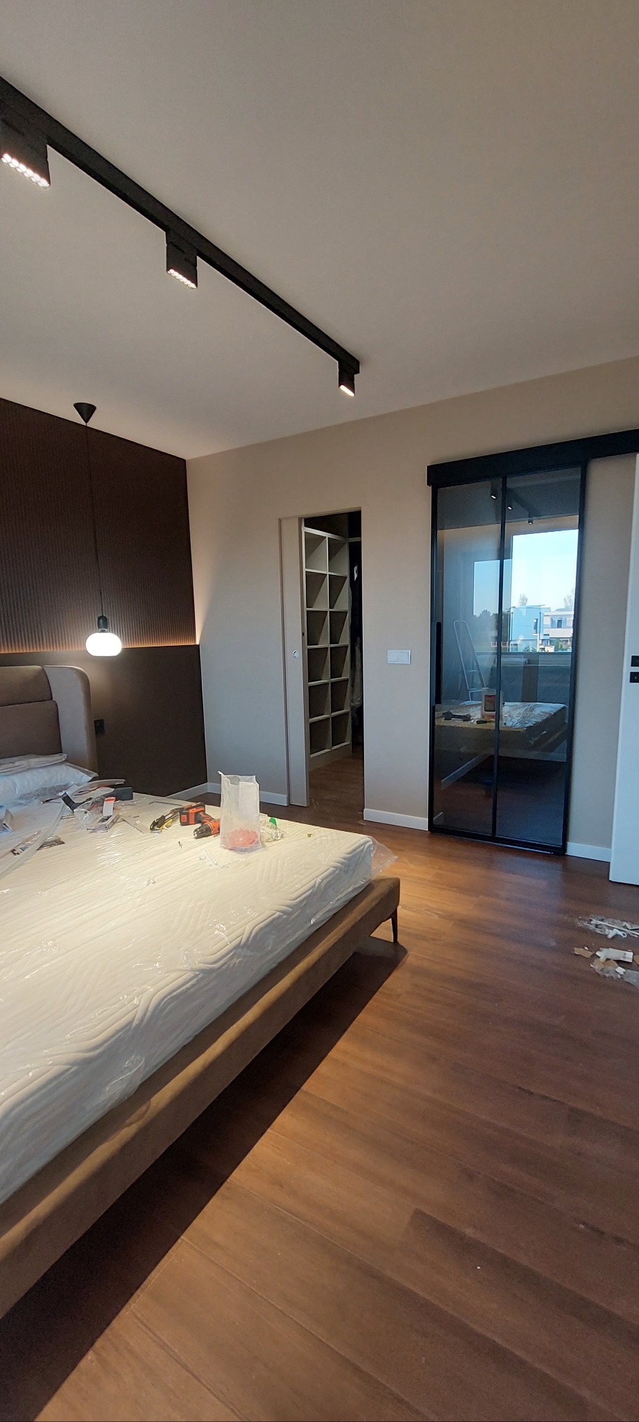 Nowoczesna sypialnia w trakcie wykańczania: drewniana podłoga, przesuwne drzwi z czarną ramą, garderoba w głębi. Na łóżku narzędzia i folia ochronna.