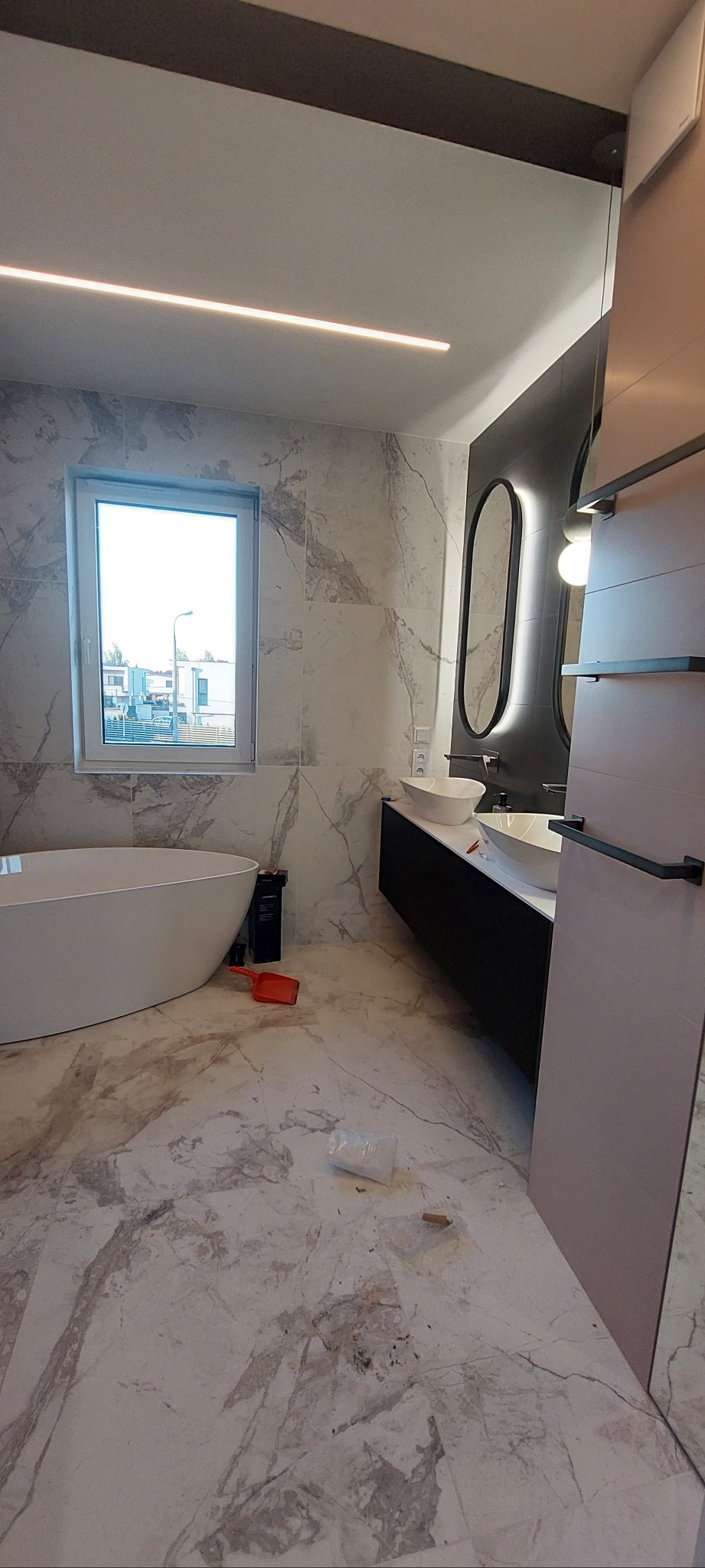 Nowoczesna łazienka z marmurowymi płytkami, wolnostojącą wanną i owalnymi lustrami z podświetleniem LED. Widoczne niedociągnięcia na podłodze.