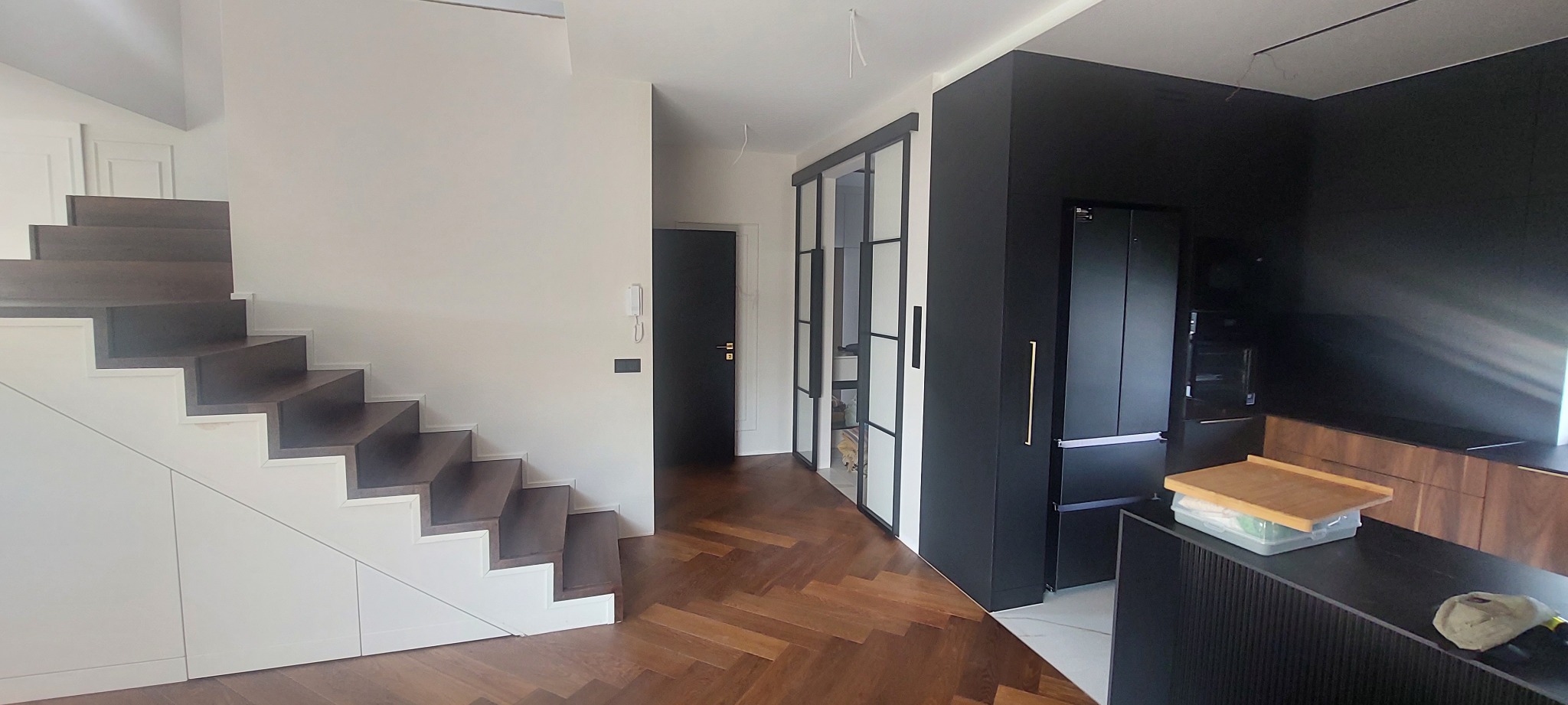 Nowoczesne wykończenie wnętrza w Oławie: drewniana podłoga w jodełkę, schody z zabudową, czarna zabudowa kuchenna i przesuwne drzwi ze szprosami.