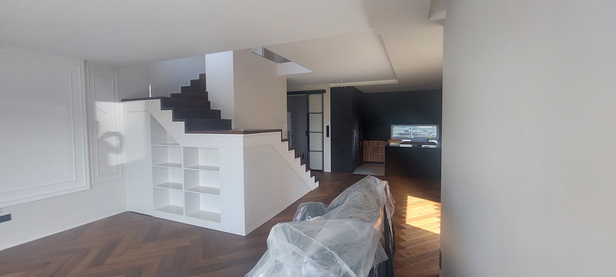 Nowoczesne wnętrze z drewnianą podłogą w jodełkę, białymi schodami z wbudowanymi półkami i czarną kuchnią w tle. Sofa zabezpieczona folią ochronną.