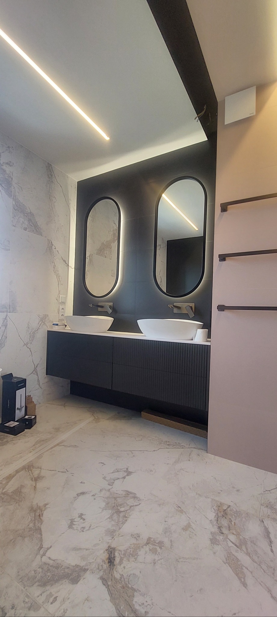 Nowoczesna łazienka z marmurową podłogą i ścianami, podwójna umywalka na szafce z ryflowanym frontem, owalne lustra z podświetleniem LED i grzejnik drabinkowy.