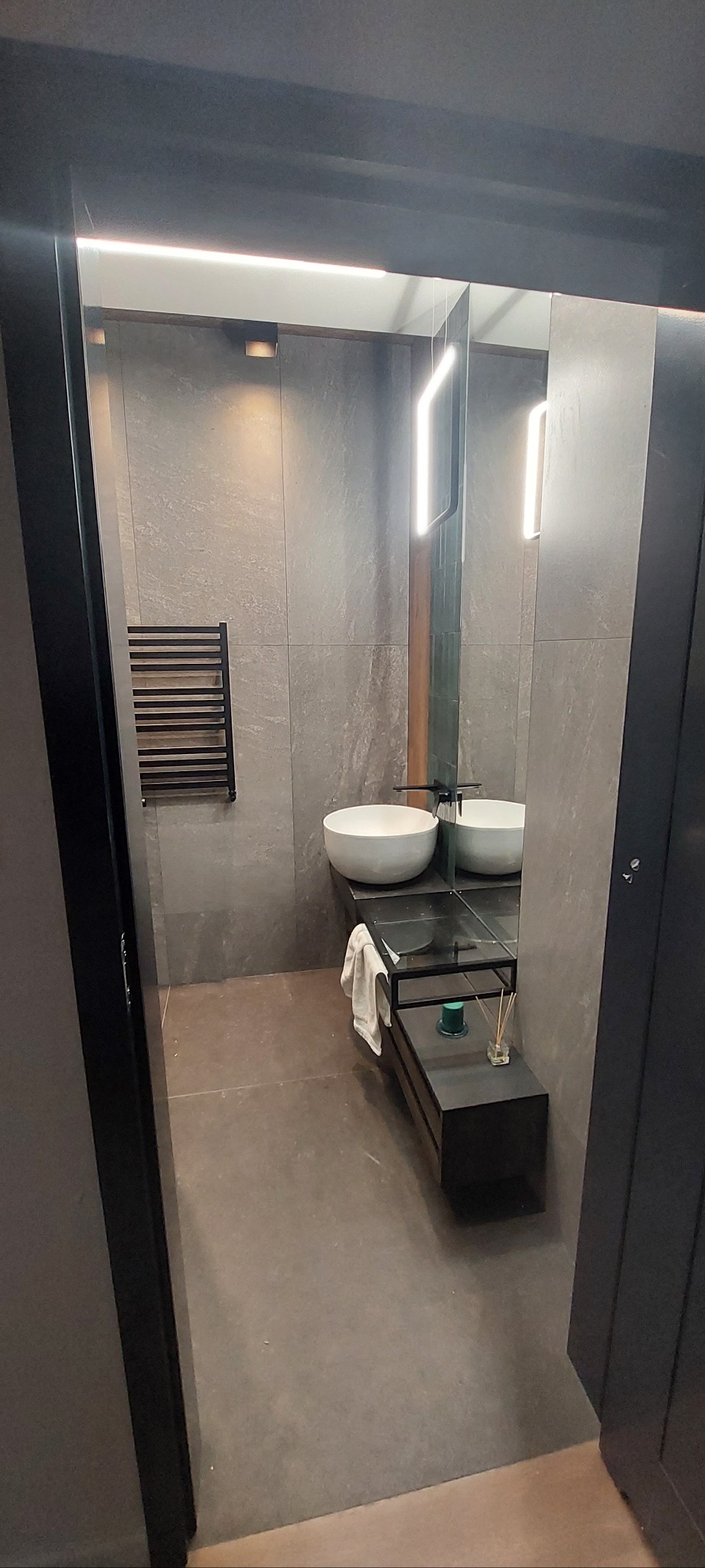 Nowoczesna łazienka w szarościach z okrągłą umywalką, podświetlanym lustrem i czarnym grzejnikiem drabinkowym. Minimalistyczny design, eleganckie wykończenie.