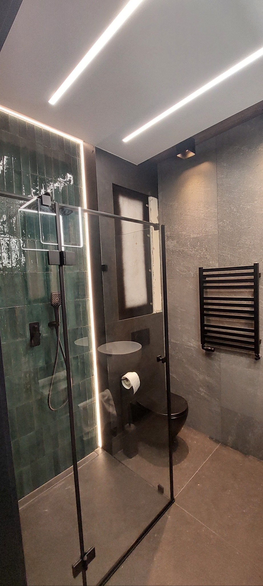 Nowoczesna łazienka z kabiną typu walk-in, zielonymi płytkami i czarną armaturą. Oświetlenie LED podkreśla minimalistyczny design. Grzejnik drabinkowy w kolorze czarnym.