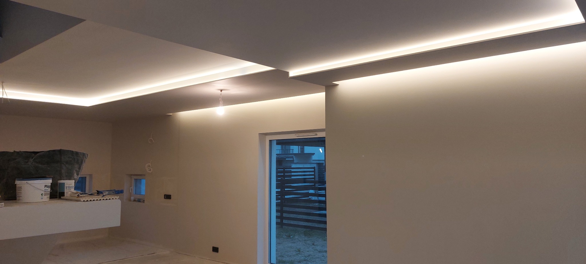 Nowoczesne oświetlenie sufitowe LED w jasnym wnętrzu. Widoczne puszki z farbą i narzędzia malarskie, co sugeruje etap wykończeniowy. Minimalistyczny design.