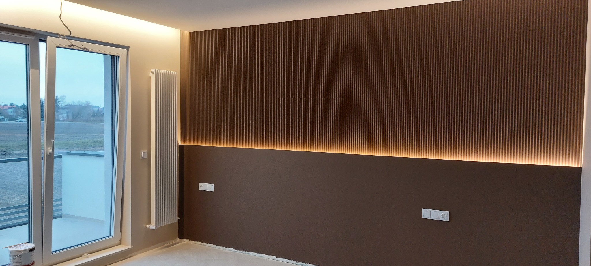 Nowoczesna ściana z brązowymi panelami i subtelnym podświetleniem LED, grzejnik, okno balkonowe. Minimalistyczny design wnętrza.