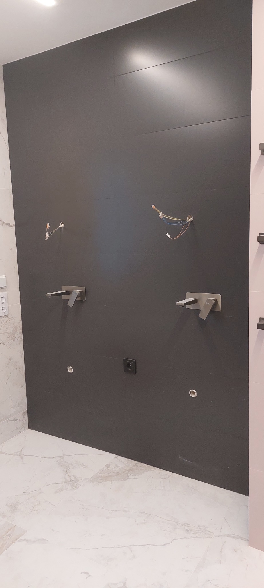 Ciemna ściana z zamontowaną armaturą łazienkową i instalacją elektryczną w trakcie prac wykończeniowych. Widoczne płytki i marmurowa podłoga.