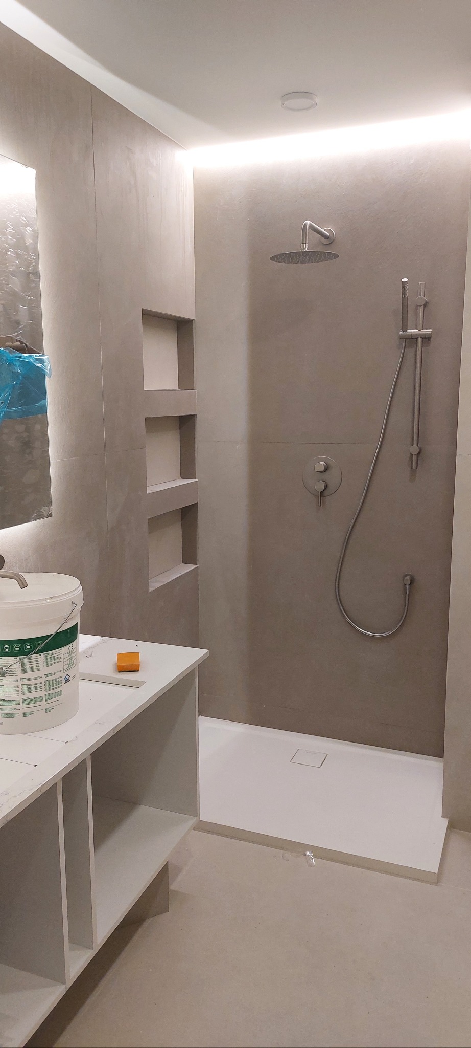 Wykończona łazienka z kabiną typu walk-in, szare płytki, wnęki w ścianie. Widoczny biały brodzik i bateria prysznicowa. Na pierwszym planie biała szafka i wiadro z farbą.