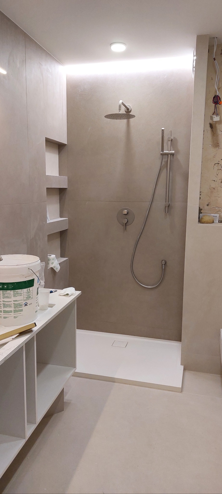 Nowoczesna kabina prysznicowa z deszczownicą i półkami w ścianie, w trakcie wykańczania. Widoczne narzędzia i materiały budowlane. Minimalistyczny design.