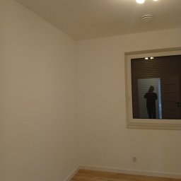 Na Gotowo Łukasz Lupa - Jasne wnętrze pokoju po remoncie: świeżo pomalowane ściany w odcieniach bieli, drewniana podłoga i okno z widoczną sylwetką osoby wewnątrz.