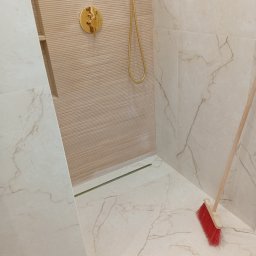 Na Gotowo Łukasz Lupa - Wykończona kabina prysznicowa z beżowymi płytkami imitującymi marmur, złotą armaturą i odpływem liniowym. Na podłodze leży czerwona zmiotka.