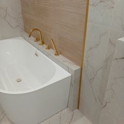 Na Gotowo Łukasz Lupa - Nowoczesna łazienka z białą wanną i złotą armaturą. Płytki imitujące marmur na podłodze i ścianach, beżowa ściana z poziomymi żłobieniami i złotym akcentem.