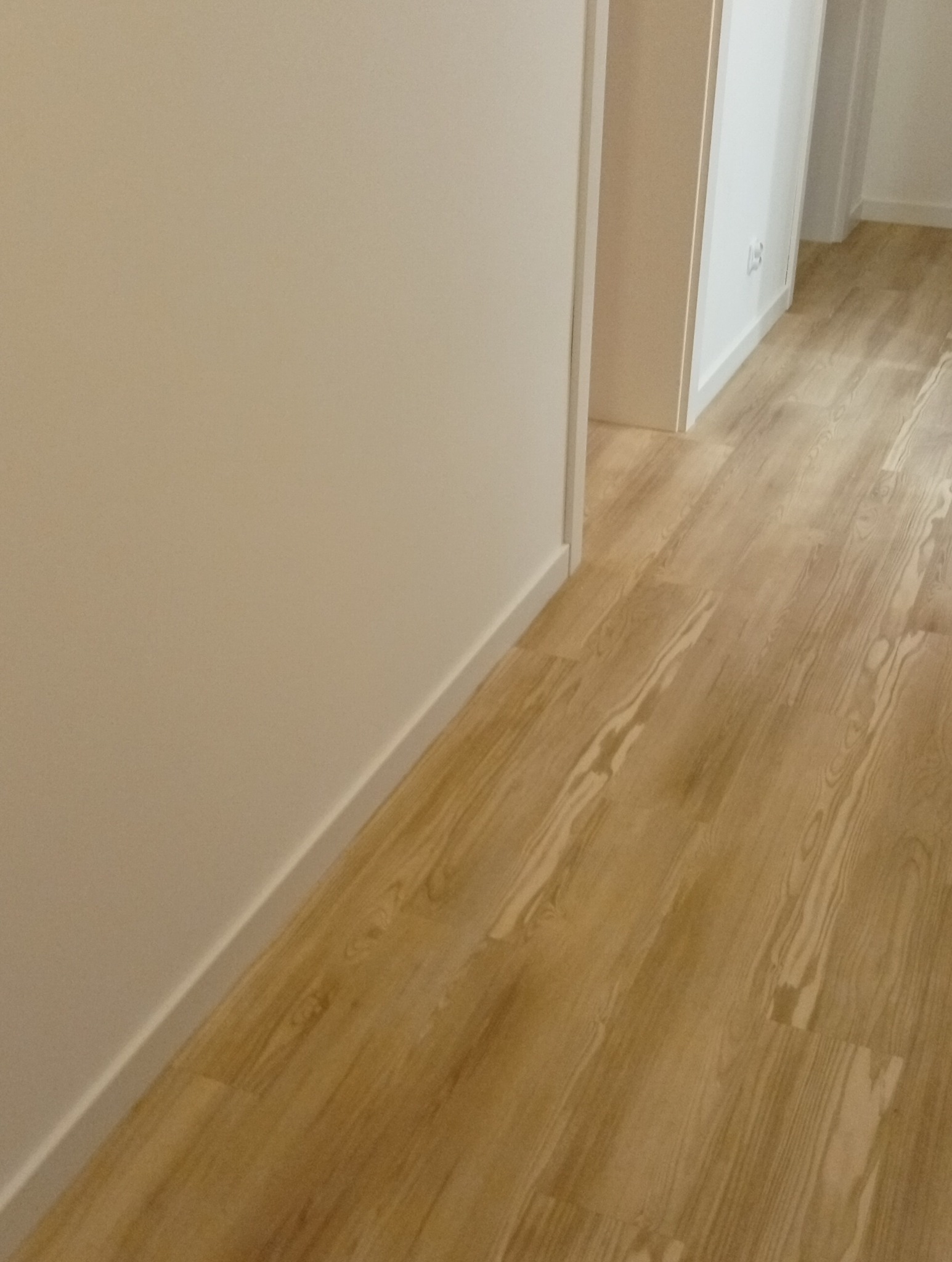 Jasne panele podłogowe ułożone wzdłuż ściany z listwą przypodłogową. Widok na korytarz z białymi drzwiami i ścianami. Neutralna kolorystyka wnętrza.