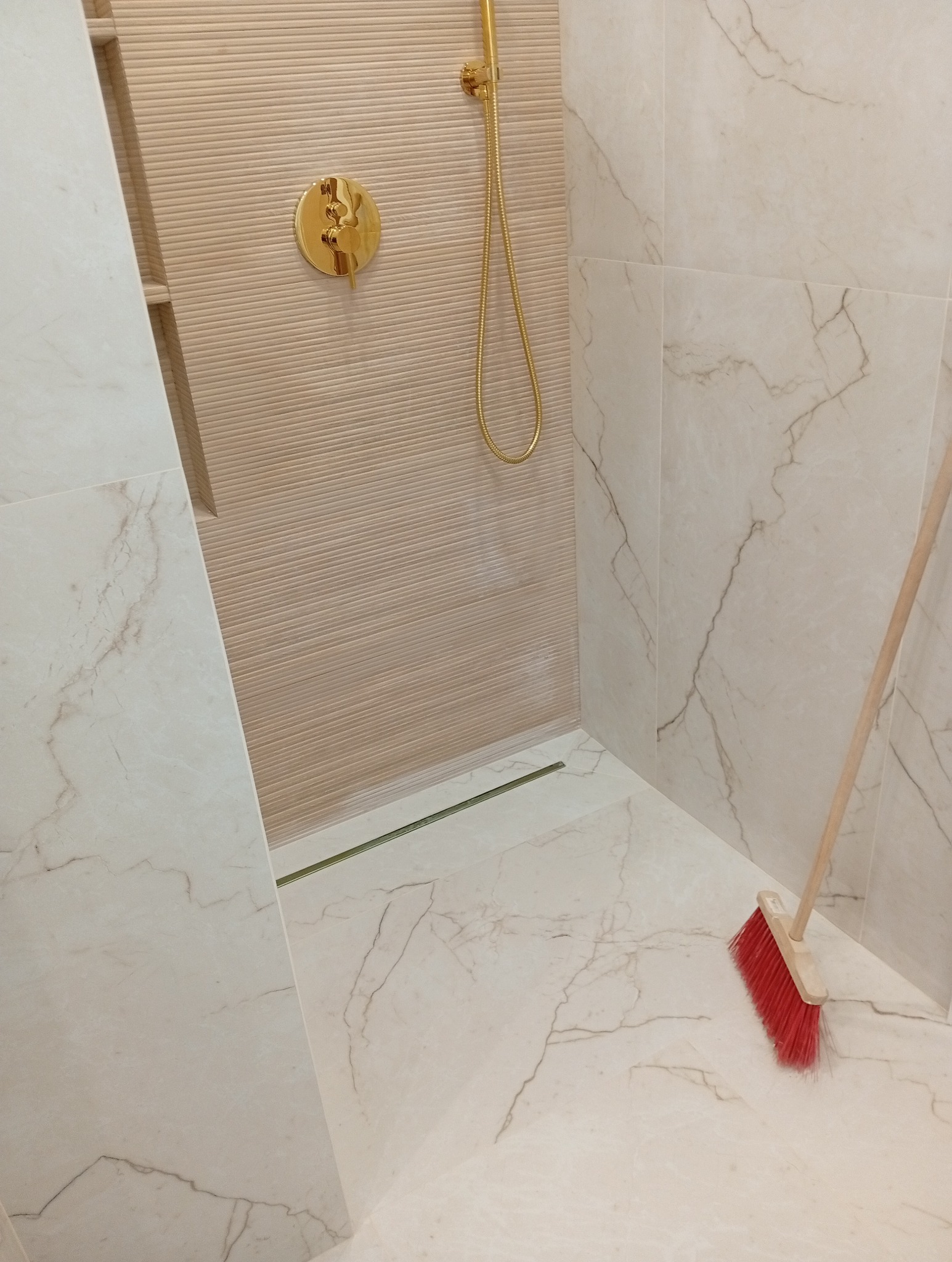 Wykończona kabina prysznicowa z beżowymi płytkami imitującymi marmur, złotą armaturą i odpływem liniowym. Na podłodze leży czerwona zmiotka.