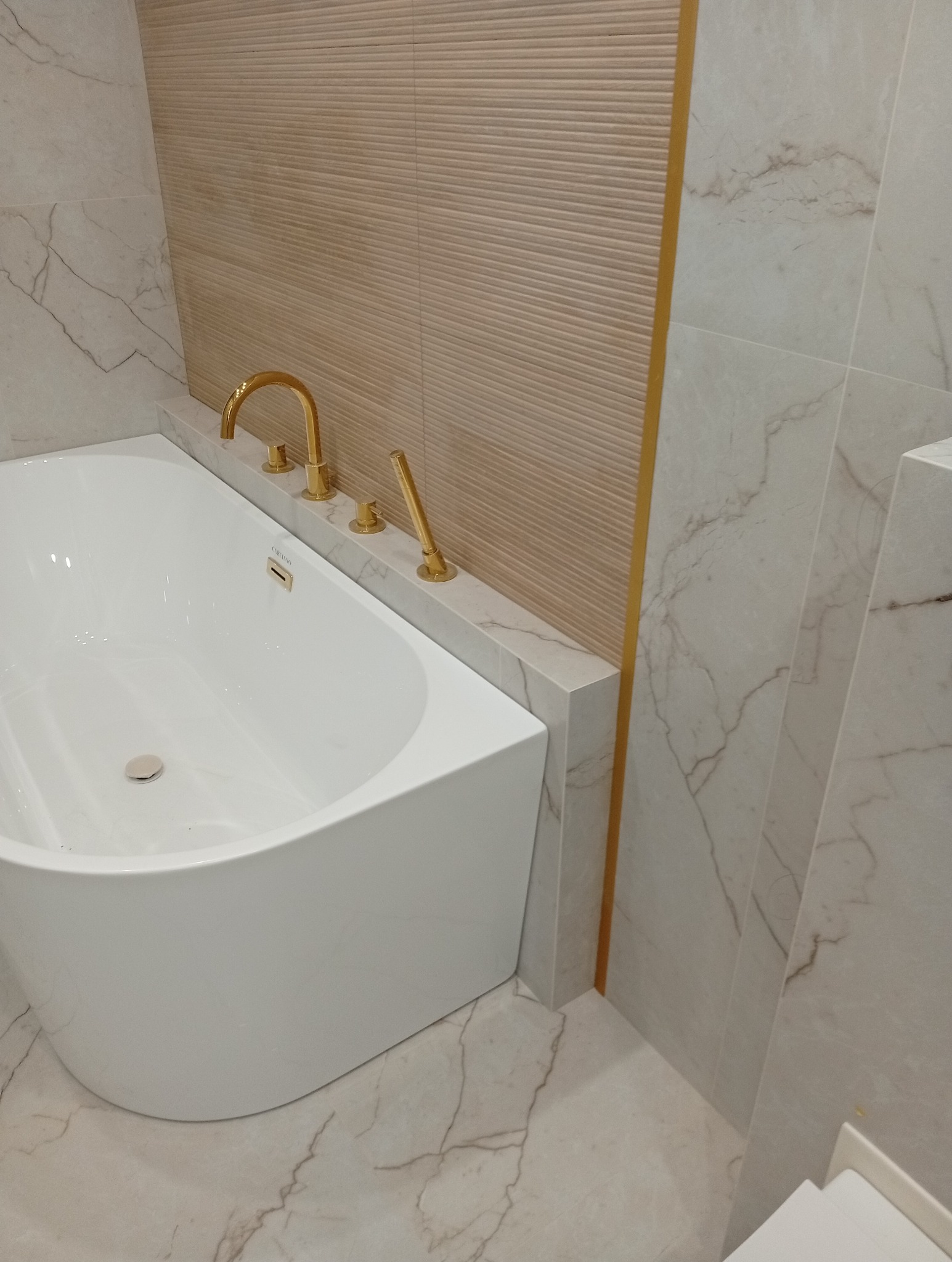 Nowoczesna łazienka z białą wanną i złotą armaturą. Płytki imitujące marmur na podłodze i ścianach, beżowa ściana z poziomymi żłobieniami i złotym akcentem.