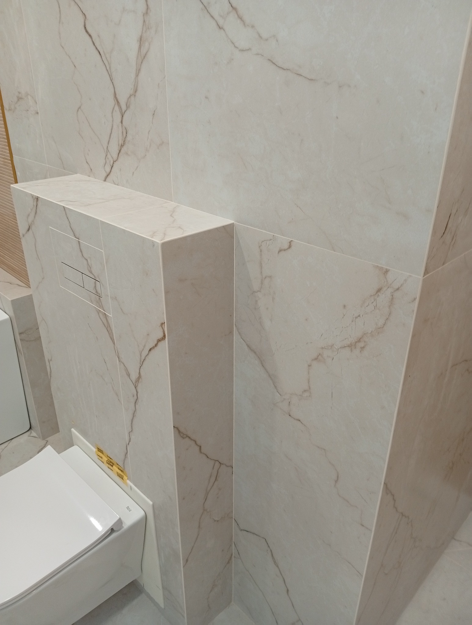 Beżowe płytki imitujące marmur w wykończonej łazience. Widoczny fragment toalety podwieszanej i zabudowa stelaża z przyciskiem spłukującym. Minimalistyczny design.
