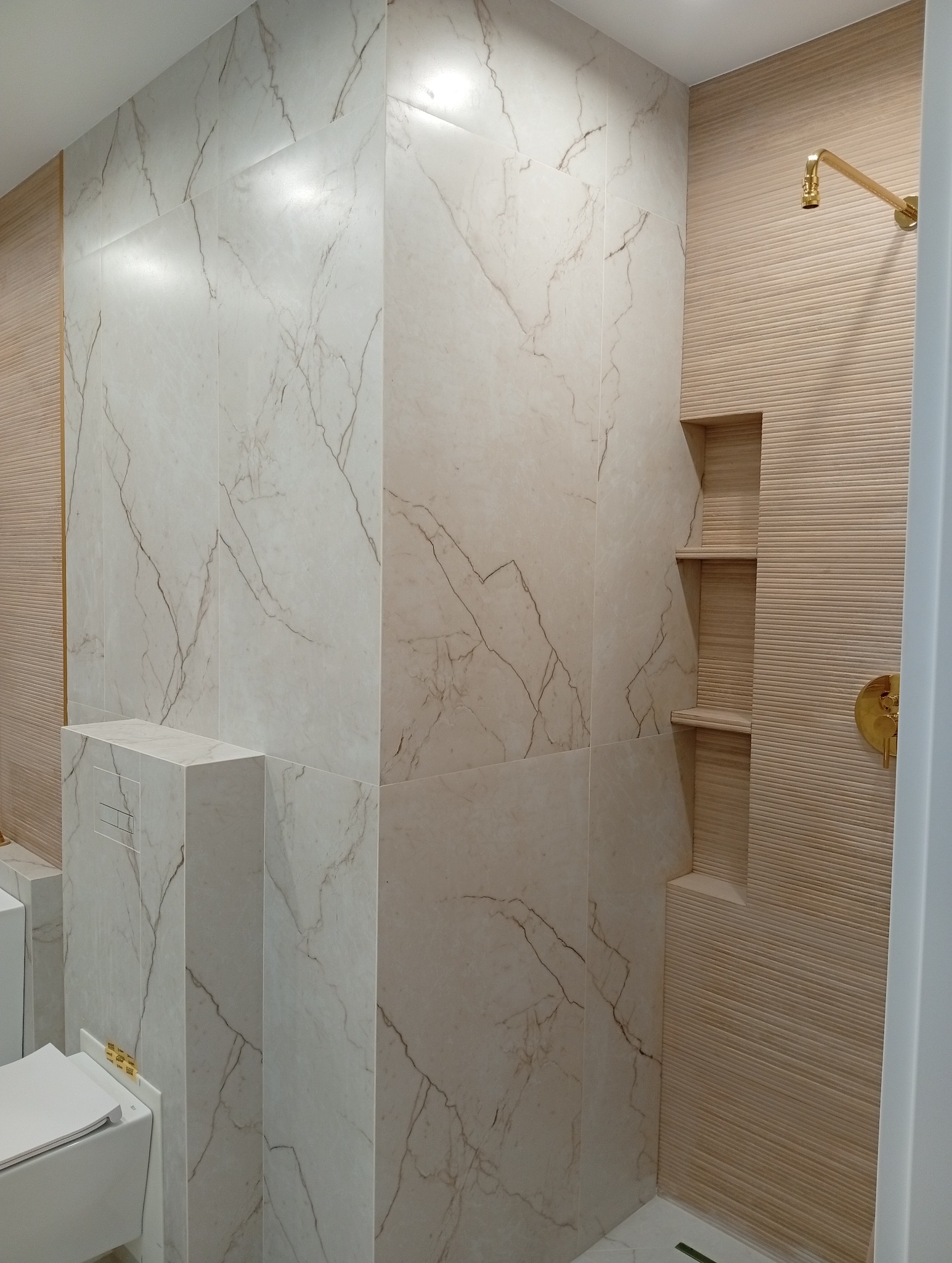 Wykończona łazienka z beżowymi płytkami imitującymi marmur i drewno. Widoczna złota armatura i wnęki w ścianie. Minimalistyczny design.