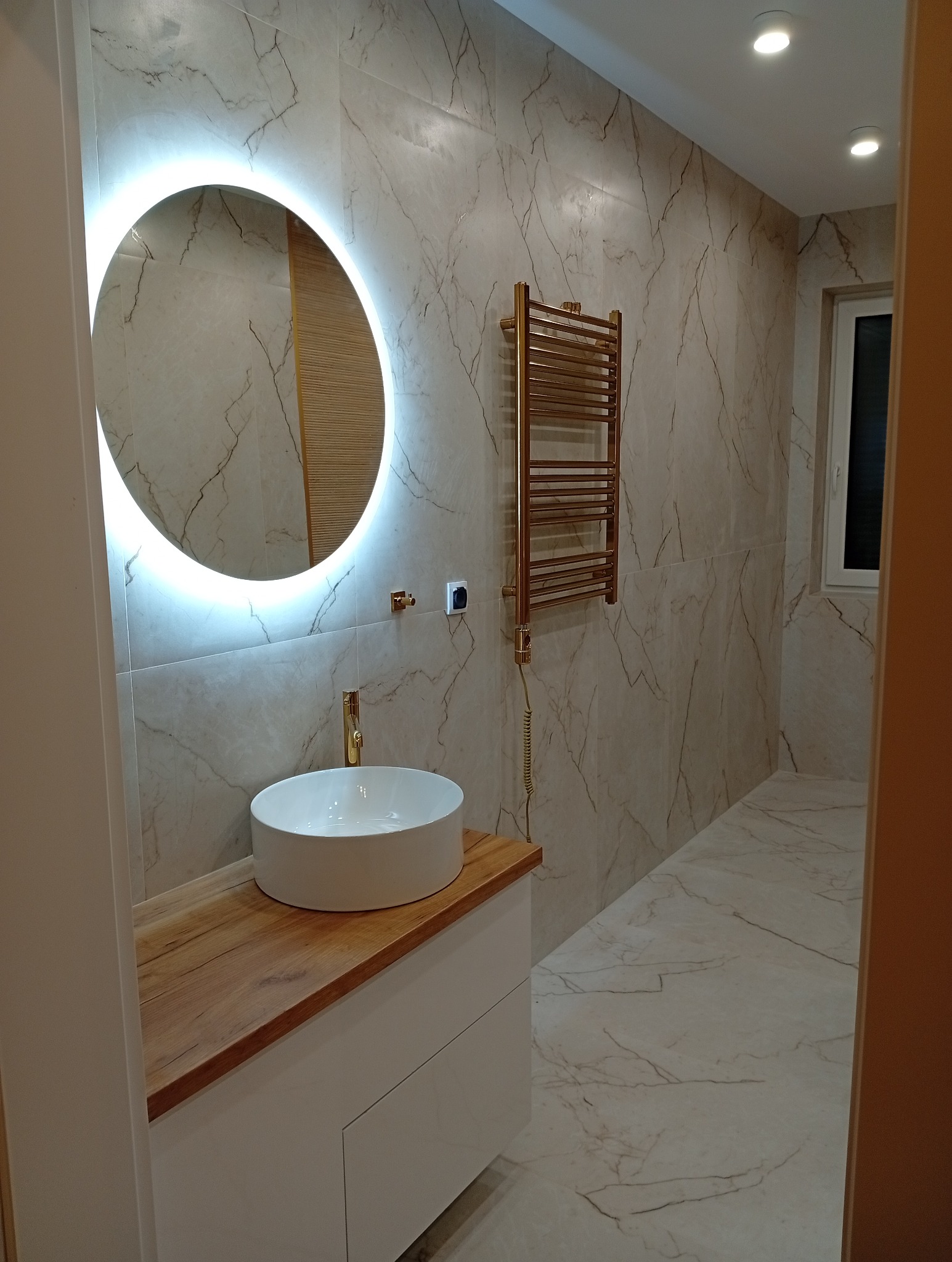 Wykończona łazienka z umywalką nablatową, okrągłym lustrem z podświetleniem LED i grzejnikiem drabinkowym w kolorze złotym. Płytki imitujące marmur na ścianach i podłodze.