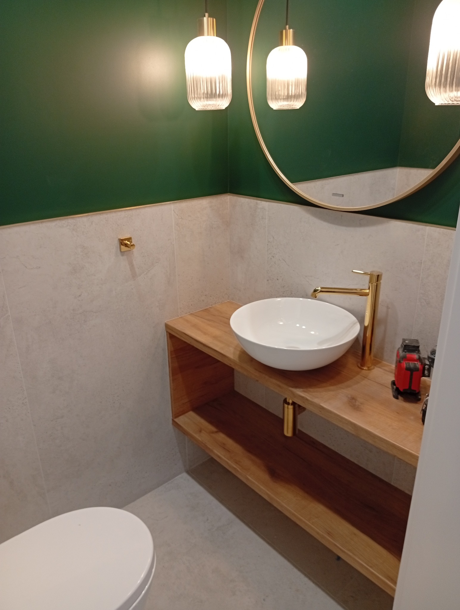Nowoczesna łazienka z umywalką nablatową, złotą armaturą i okrągłym lustrem. Zielona ściana i drewniany blat dodają elegancji. Widoczny laser krzyżowy.