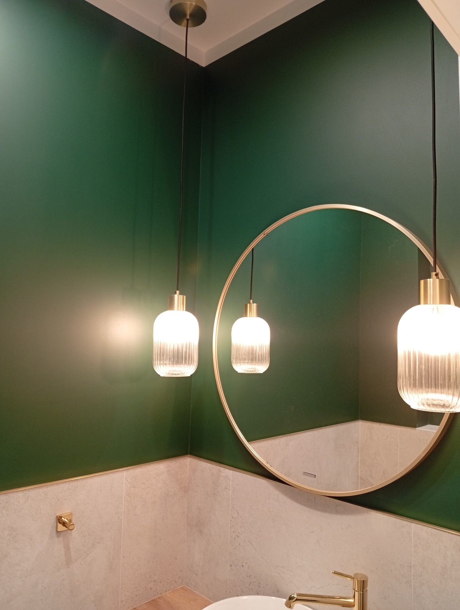 Wykończona łazienka z zieloną ścianą, okrągłym lustrem w złotej ramie i dwoma wiszącymi lampami z ryflowanego szkła. Złota armatura i listwy dekoracyjne.