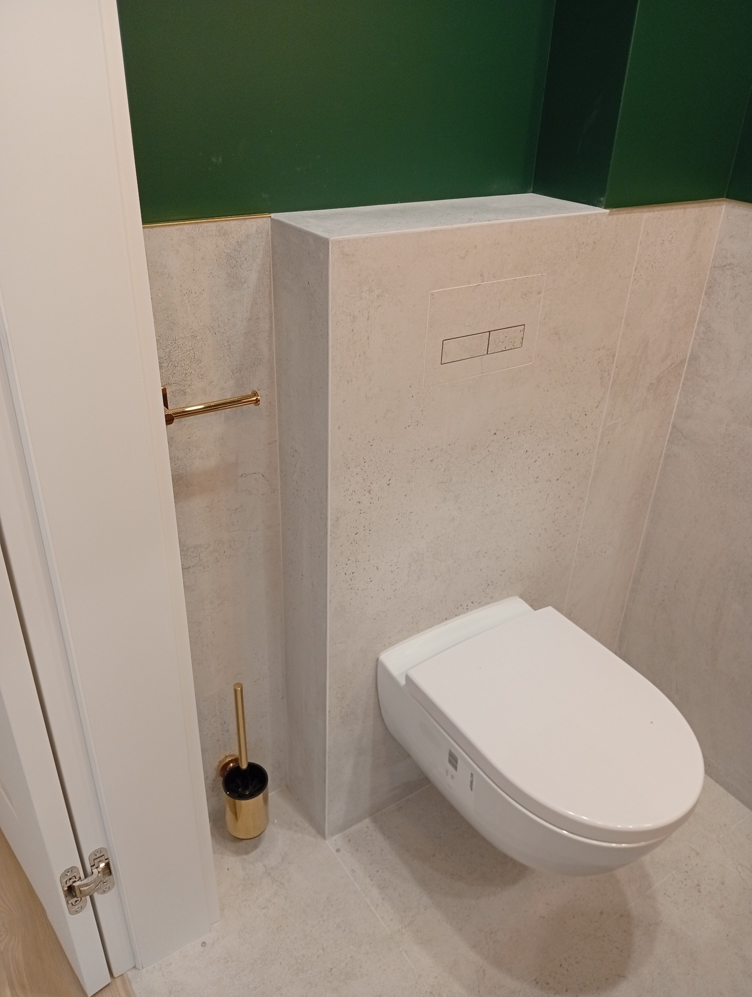 Nowoczesna toaleta podwieszana z beżowymi płytkami i złotymi akcentami, w tym uchwyt na papier i szczotka do WC. Górna część ściany pomalowana na ciemnozielono.