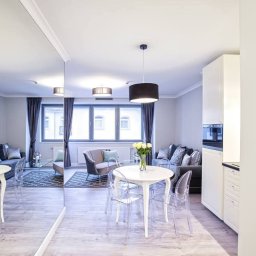 Błysk IIblask-Elzbieta Basty - Jasne wnętrze mieszkania po sprzątaniu: biały stół z kwiatami, przezroczyste krzesła, szara sofa z poduszkami, okno z zasłonami, odbicie w lustrze, minimalistyczny design.