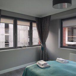 Błysk IIblask-Elzbieta Basty - Jasne wnętrze sypialni z turkusową narzutą na łóżku, szarymi zasłonami z chwostami i widokiem na budowę za oknem. Minimalistyczny wystrój, stonowana kolorystyka.