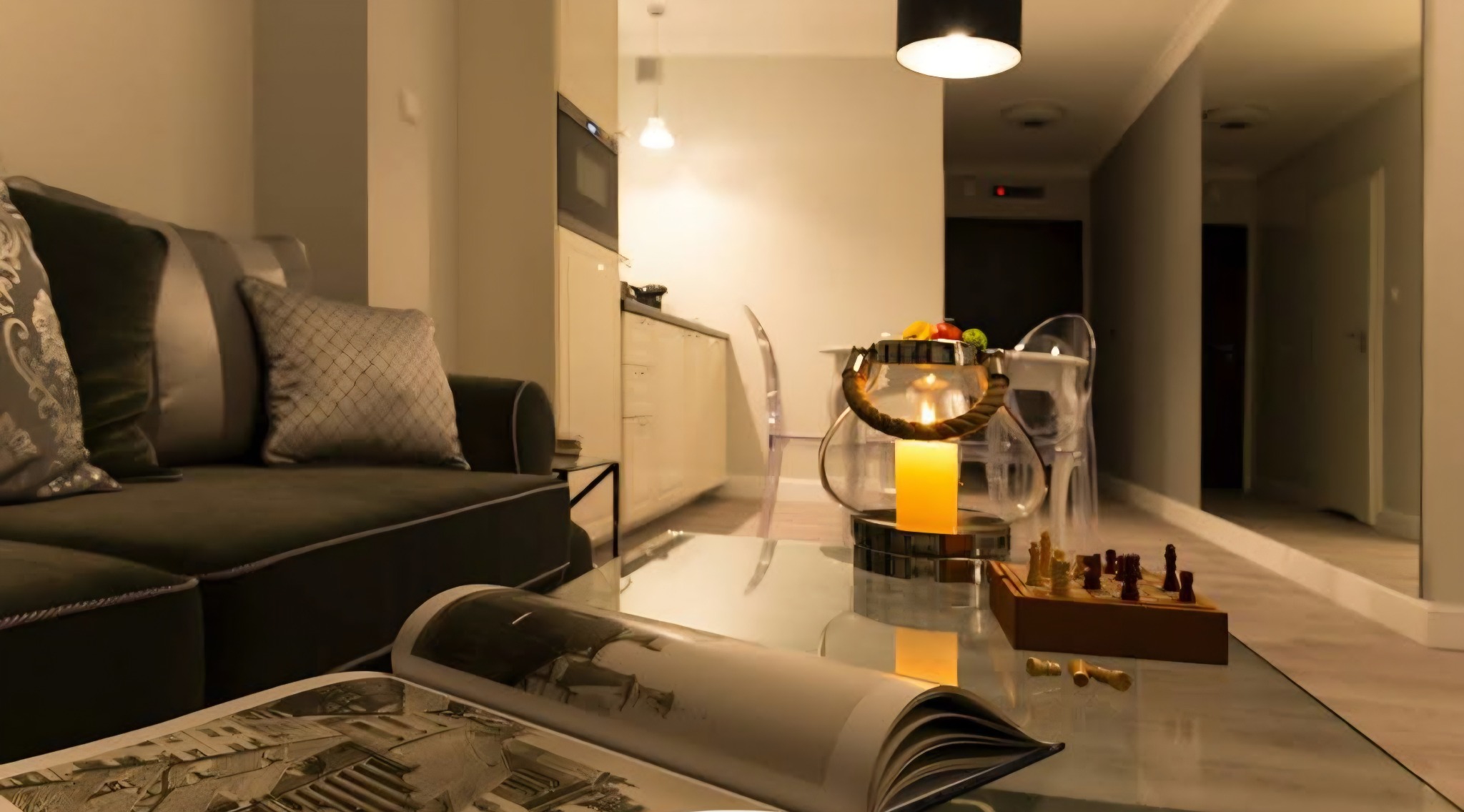Elegancki salon z szarą sofą, książką na szklanym stoliku, świecą w lampionie i szachami. W tle aneks kuchenny i lustro powiększające przestrzeń.