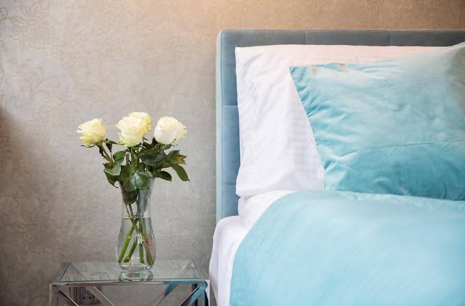 Ustawiony bukiet białych róż w szklanym wazonie na stoliku nocnym obok łóżka z błękitną pościelą i zagłówkiem. Ściana z delikatnym wzorem w tle.