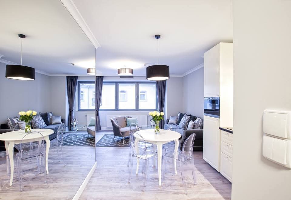 Jasne wnętrze mieszkania po sprzątaniu: biały stół z kwiatami, przezroczyste krzesła, szara sofa z poduszkami, okno z zasłonami, odbicie w lustrze, minimalistyczny design.