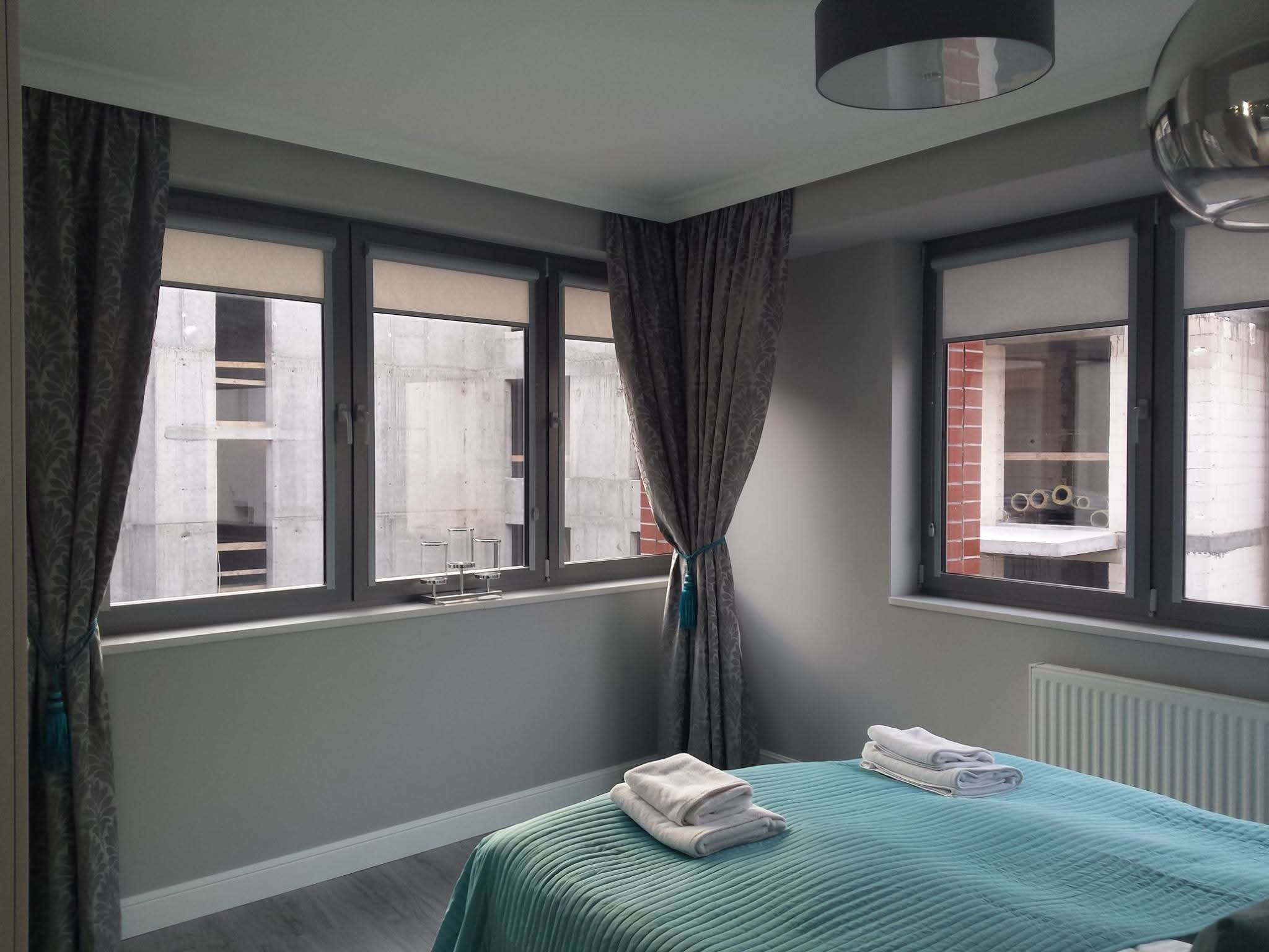 Jasne wnętrze sypialni z turkusową narzutą na łóżku, szarymi zasłonami z chwostami i widokiem na budowę za oknem. Minimalistyczny wystrój, stonowana kolorystyka.