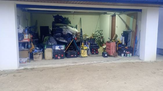 Zagracony garaż z narzędziami, sprzętem i stertą przedmiotów, czekający na opróżnienie. Widoczne skrzynki narzędziowe, rowery i inne przedmioty.