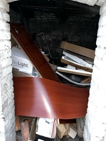 Stare meble i kartony upchnięte w ciemnym, ceglanym pomieszczeniu. Widoczne elementy wyposażenia wnętrz i resztki po remoncie. Opróżnianie piwnicy.