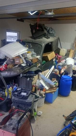 Zagracony garaż z elementami starego samochodu pod stertą przedmiotów. Widoczne narzędzia, pojemniki i różne przedmioty codziennego użytku.