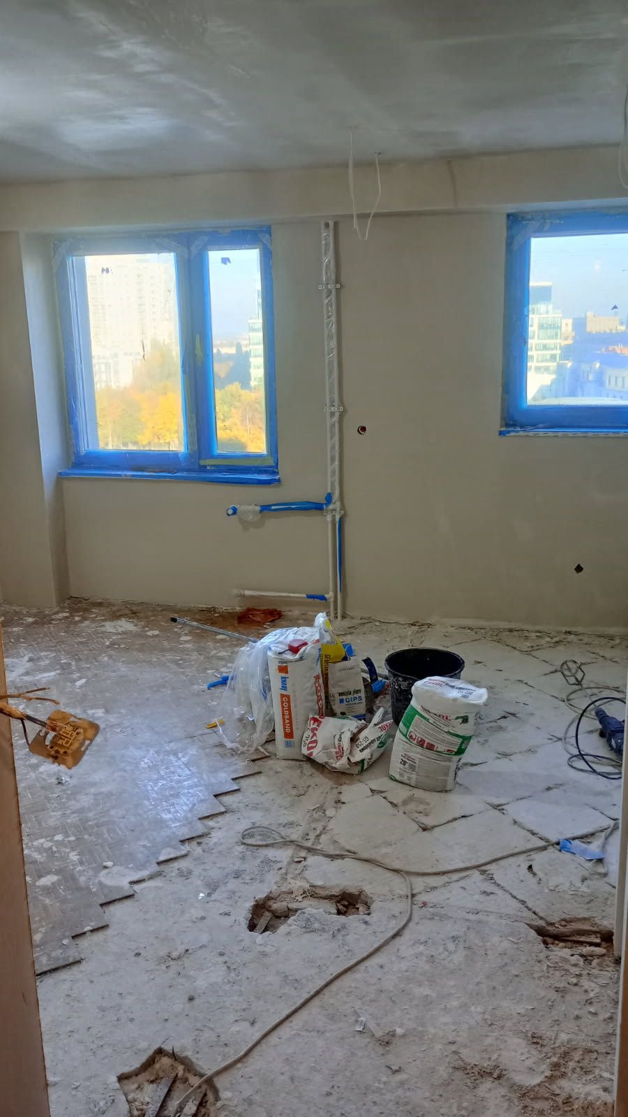Remont pokoju: zerwane płytki podłogowe, worki z gipsem, okna zabezpieczone folią. Widok na miasto przez okna. Przygotowanie do dalszych prac wykończeniowych.
