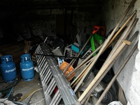 Zagracone pomieszczenie z butlami gazowymi, drabiną i narzędziami ogrodniczymi. Nieuporządkowany składzik z różnorodnymi przedmiotami w ciemnym wnętrzu.