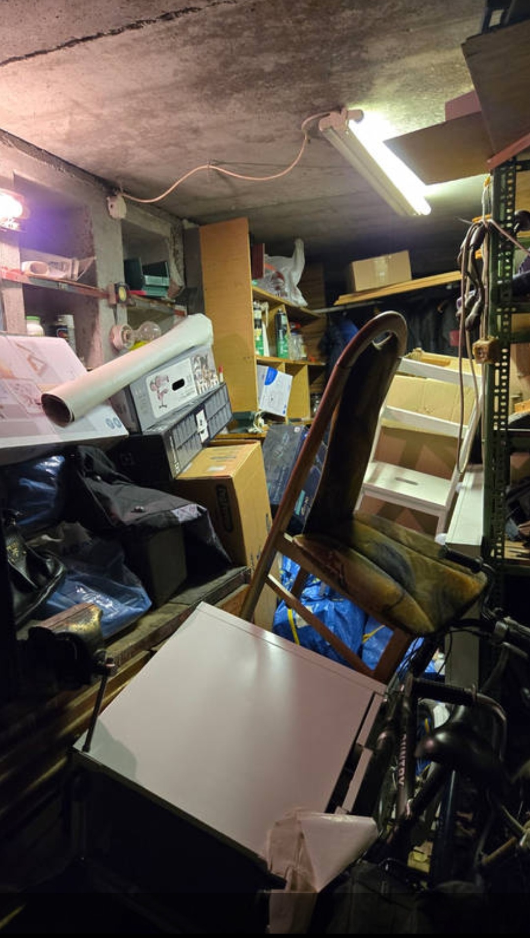 Zagracone pomieszczenie piwniczne z różnymi przedmiotami, w tym krzesłem, kartonami, rowerem i workami. Słabe oświetlenie podkreśla chaos i potrzebę posprzątania.