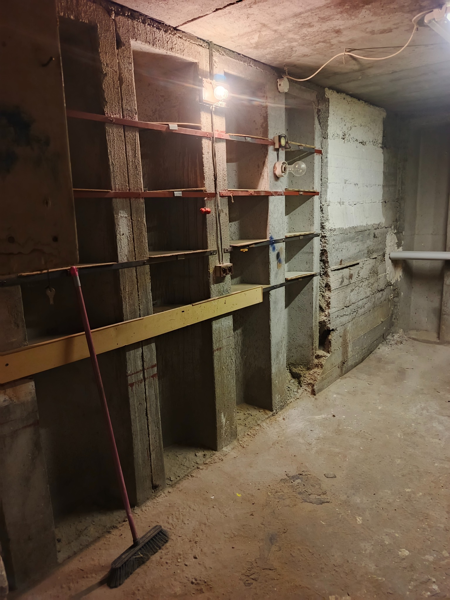 Piwnica w trakcie opróżniania: betonowe ściany z wnękami, prowizoryczne półki, widoczna instalacja elektryczna i stojąca miotła. Surowy, industrialny klimat.