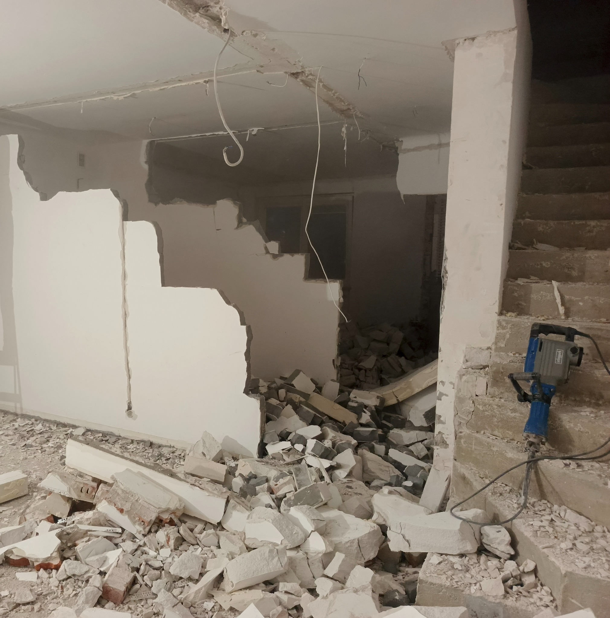 Wnętrze w trakcie rozbiórki: gruz na podłodze, uszkodzona ściana z widocznymi przewodami i schody z młotem pneumatycznym. Prace rozbiórkowe w pomieszczeniu.