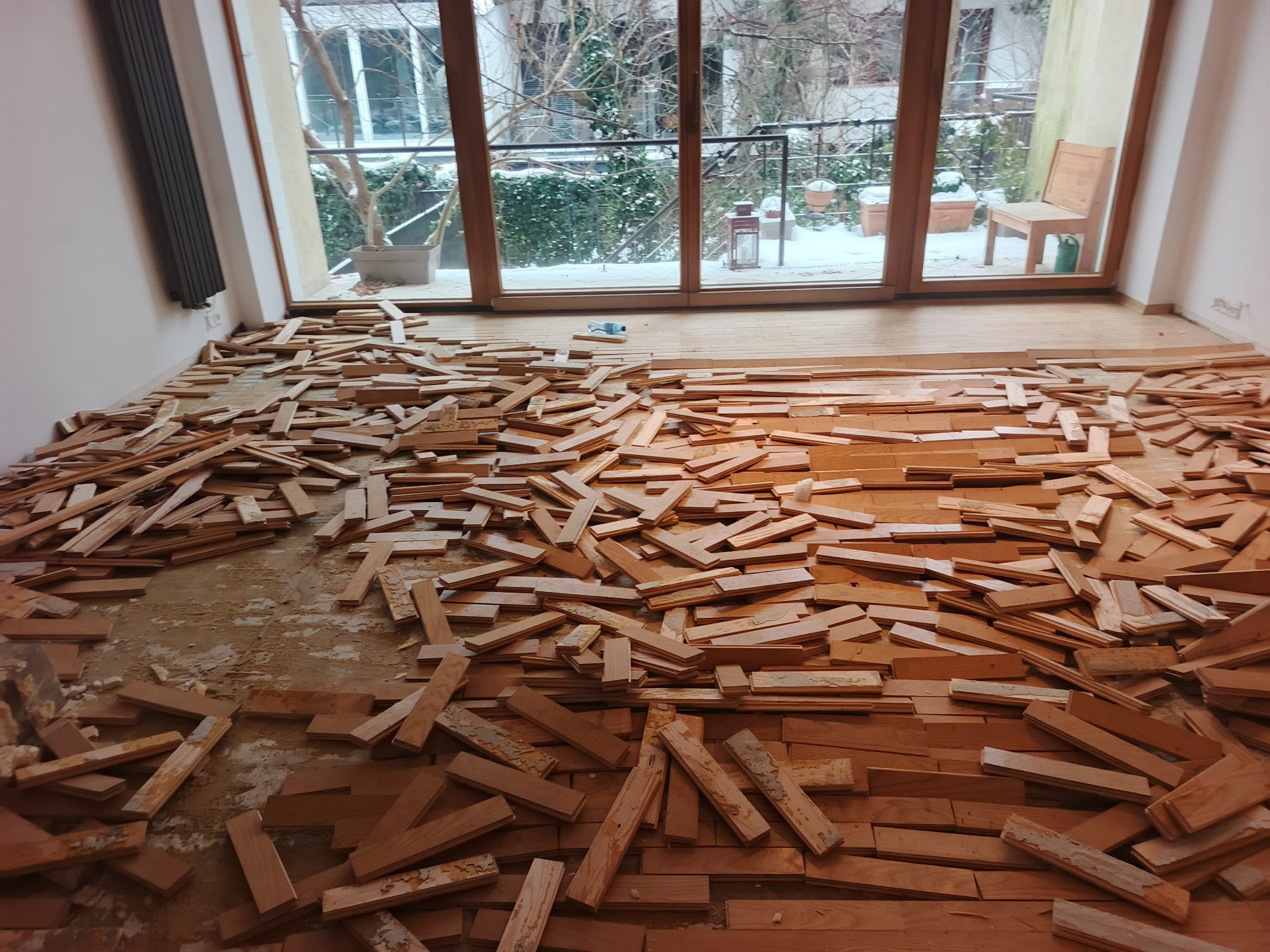 Demontaż parkietu w pokoju z widokiem na balkon. Fragmenty drewnianej podłogi porozrzucane na podłożu. Widoczne ślady kleju i starej podłogi.
