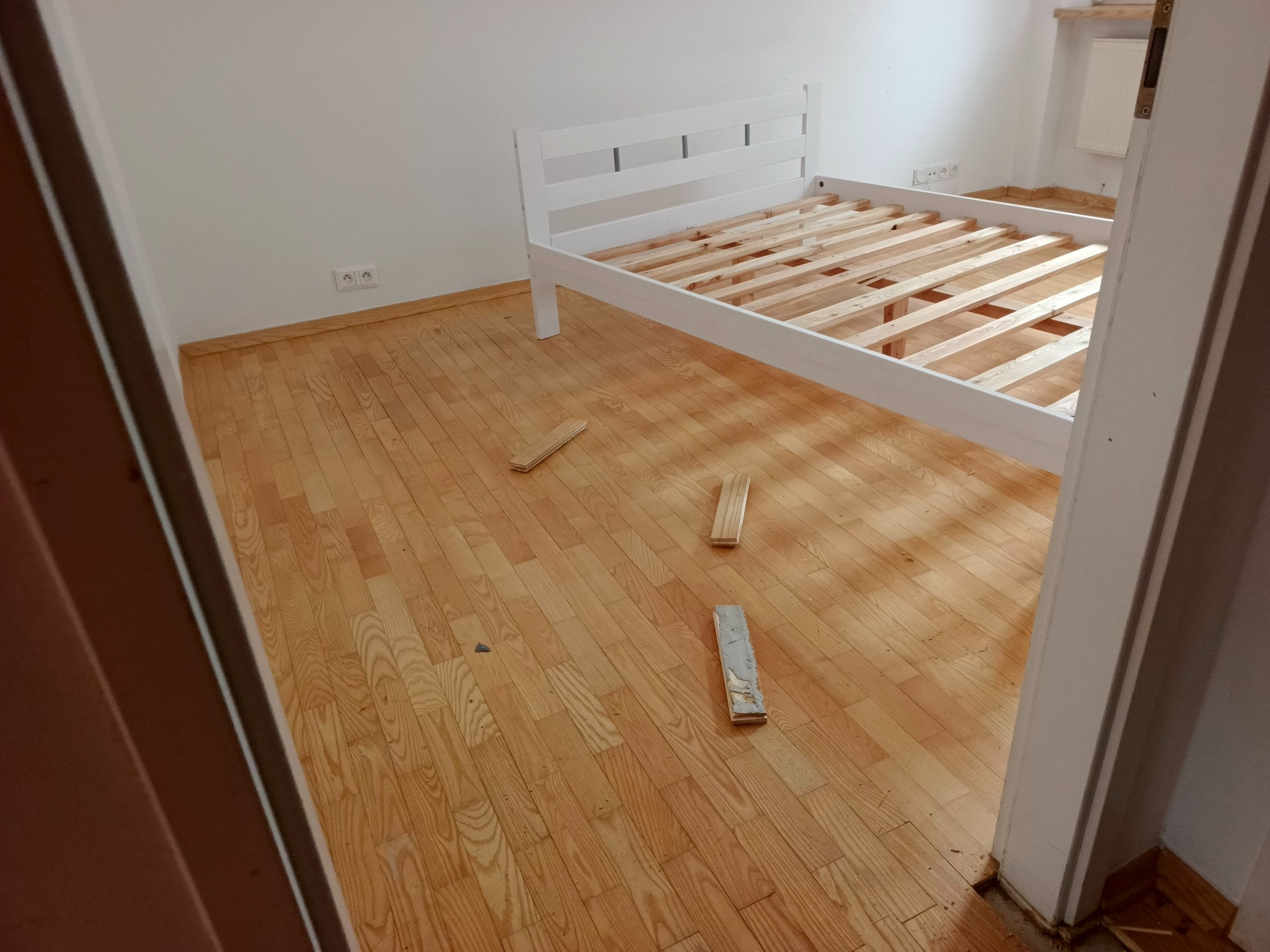 Demontaż białego łóżka z drewnianym stelażem na parkiecie. Widoczne elementy łóżka na podłodze. Ujęcie z perspektywy wejścia do pokoju.