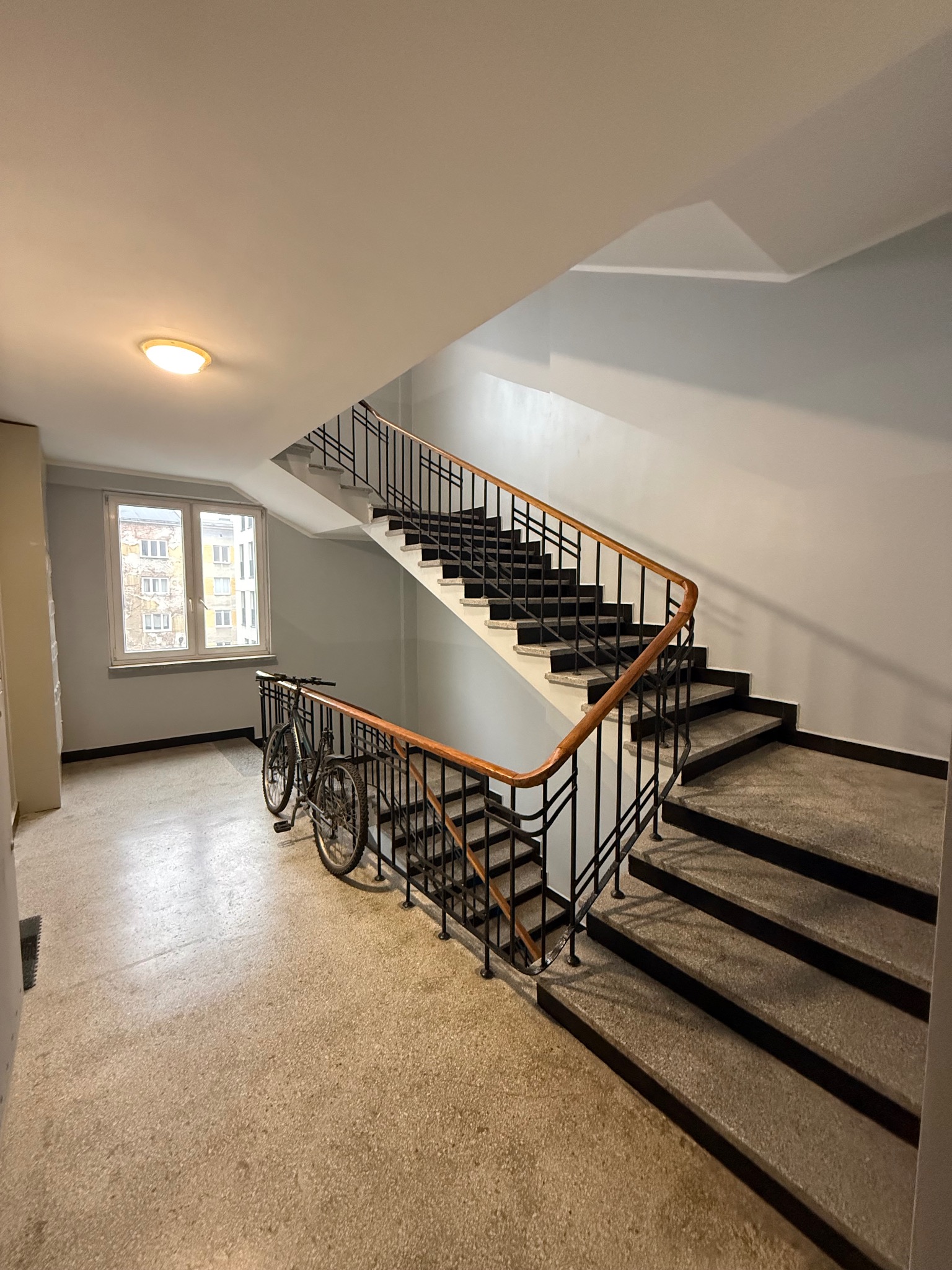 Klatka schodowa w bloku z rowerem opartym o barierkę. Widok na okno z widokiem na inne budynki. Terrazzo na podłodze i schodach. Oświetlenie sufitowe.