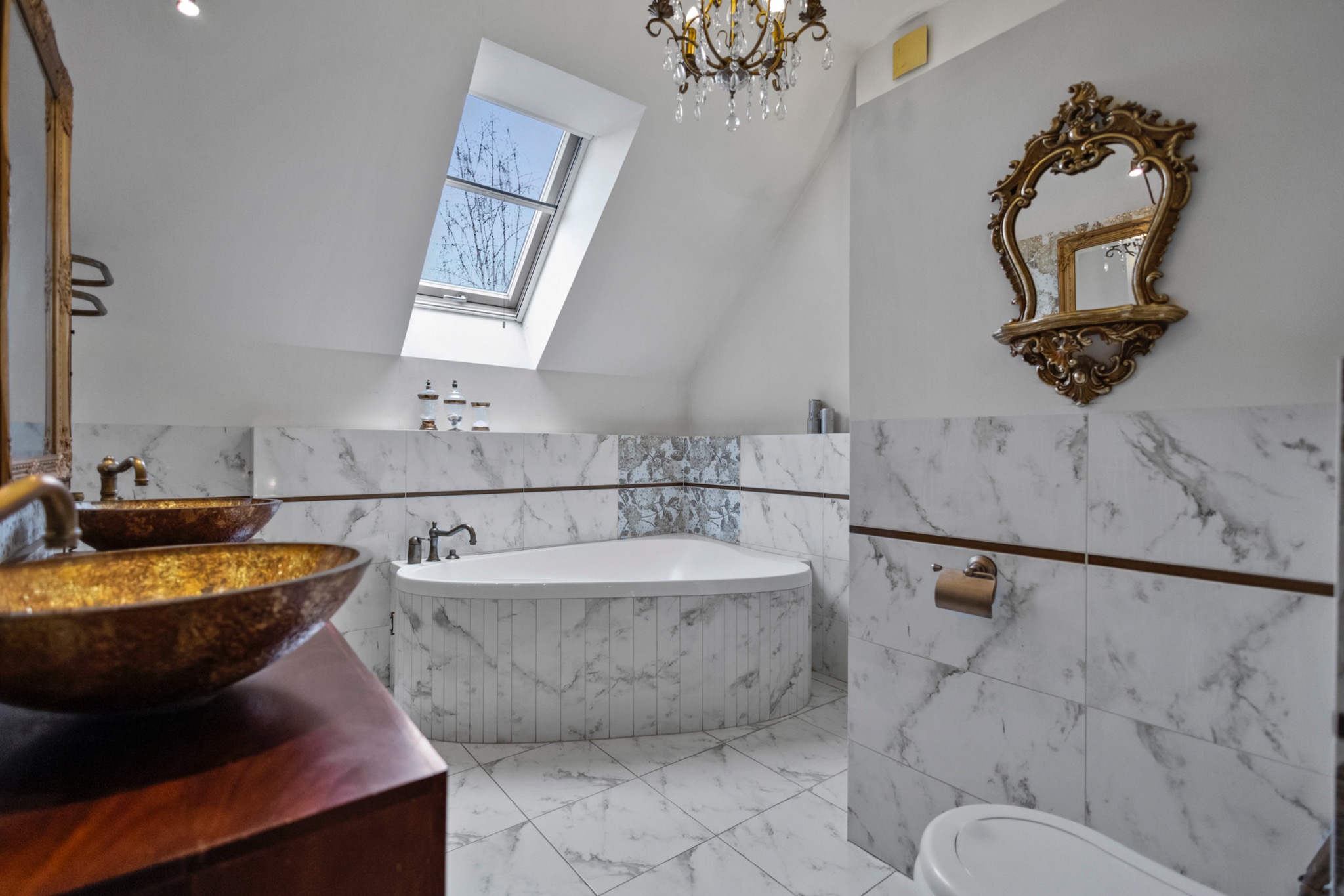 Elegancka łazienka z marmurowymi płytkami, wolnostojącą wanną narożną i złotymi akcentami, w tym lustro i umywalki, oświetlona naturalnym światłem z okna dachowego.