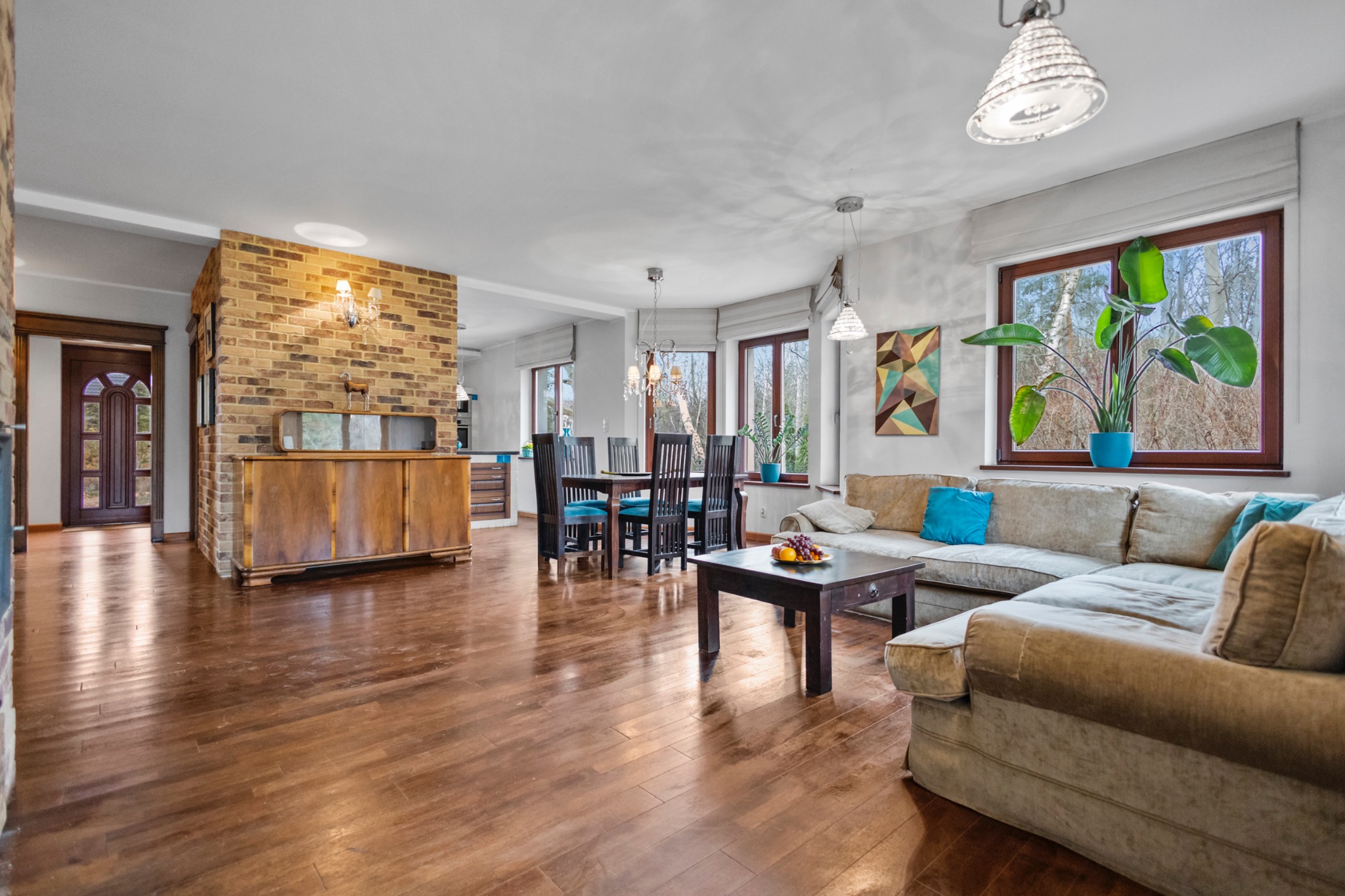 Jasne wnętrze domu z drewnianą podłogą, ceglaną ścianą i wygodną sofą. Widok na jadalnię i kuchnię. Roślina w niebieskiej doniczce na parapecie.