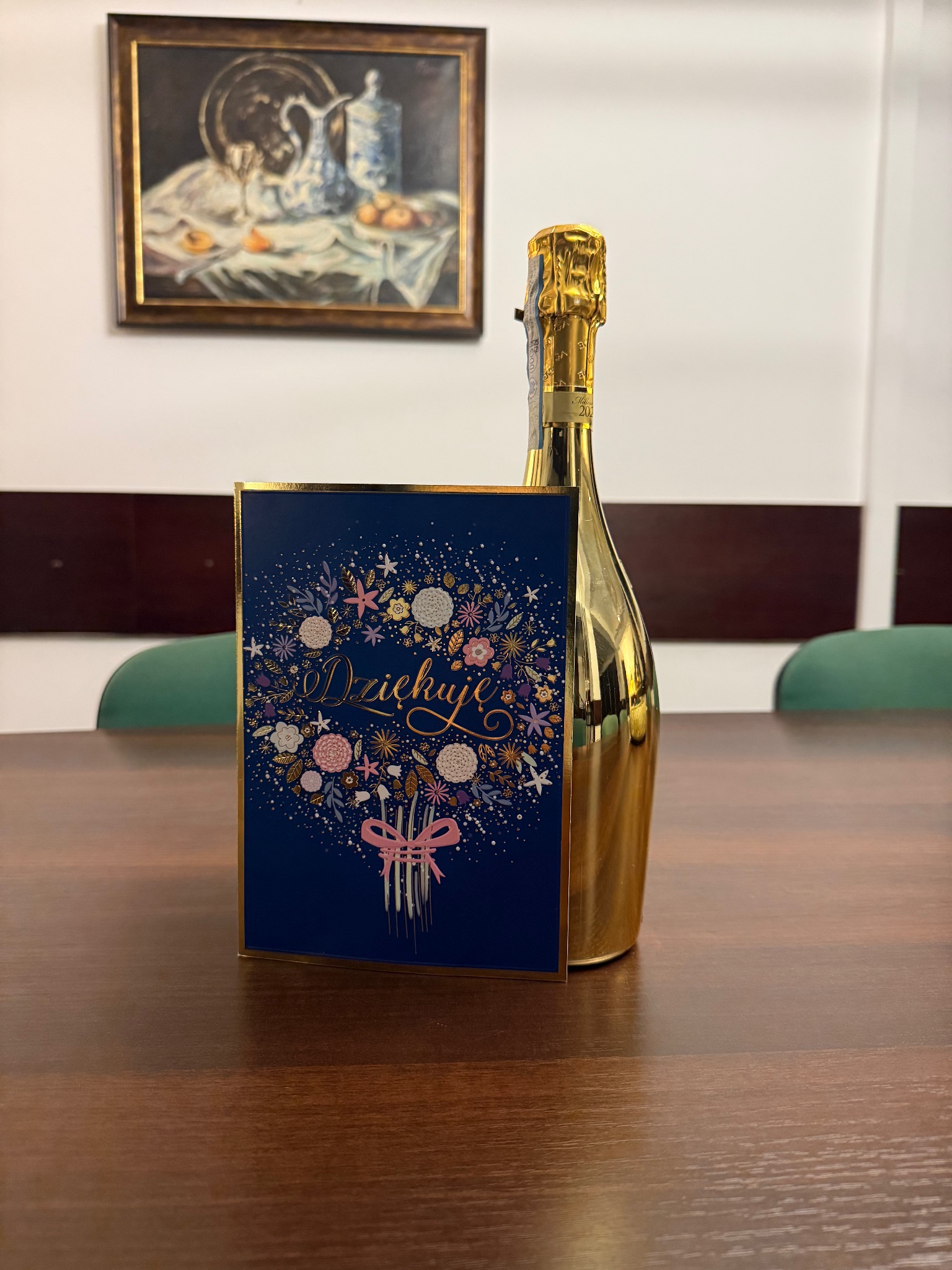 Złota butelka wina musującego i kartka z napisem 'Dziękuję' na drewnianym stole, w tle obraz z martwą naturą. Podziękowanie po transakcji.