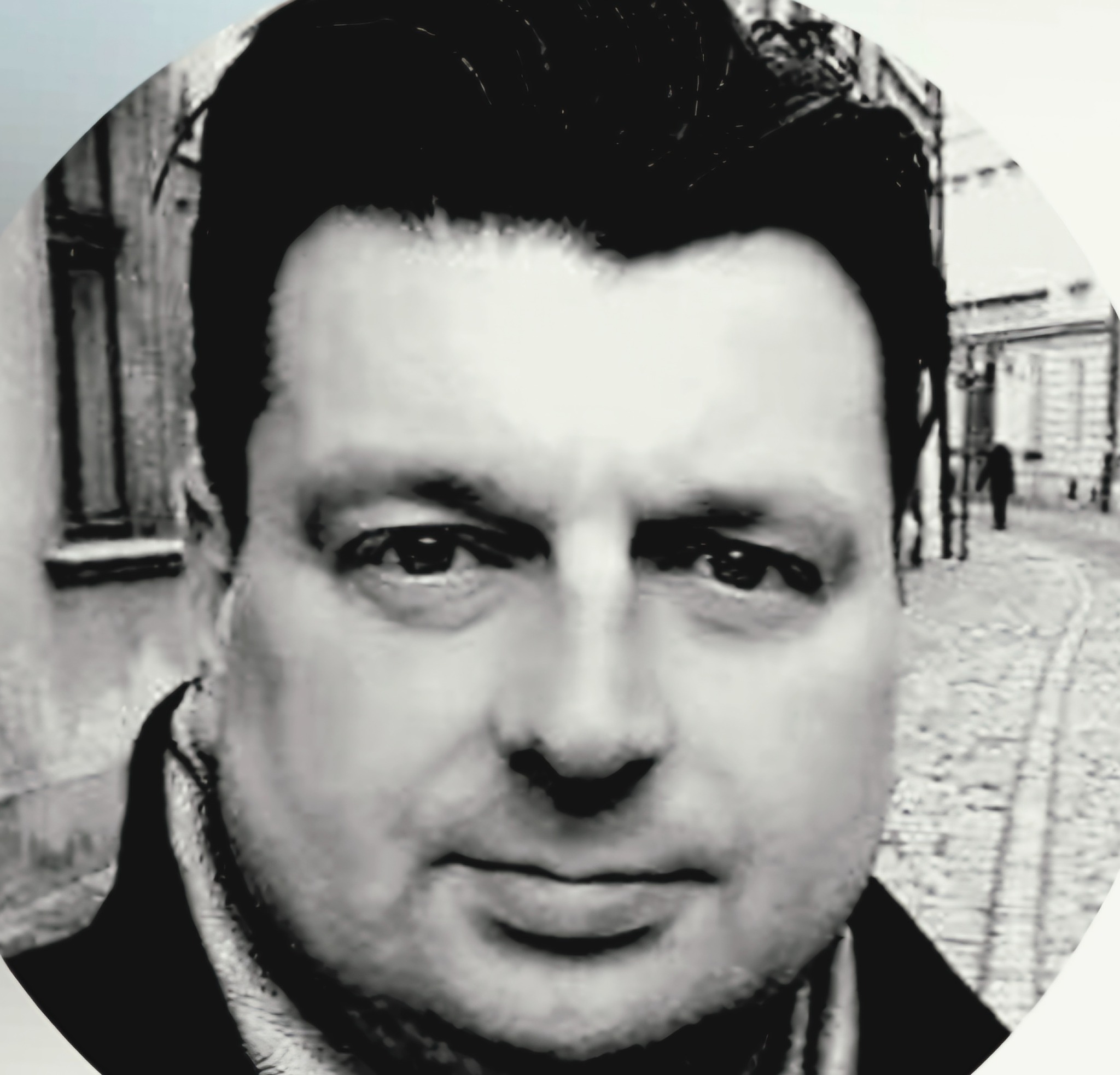 Czarno-biały portret mężczyzny na tle uliczki z brukowaną nawierzchnią i budynkami. Ujęcie w okręgu, stylizowane na retro.