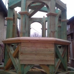 Drew-Styl - Surowa, drewniana konstrukcja altany z łukowymi przejściami, pomalowana na zielono, w trakcie budowy na tle zabudowań.