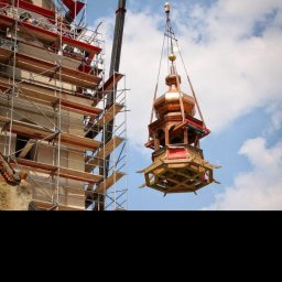 Drew-Styl - Montaż kopuły wieży kościelnej za pomocą dźwigu. Na pierwszym planie kopuła z miedzianym dachem i drewnianą konstrukcją. W tle rusztowania i fragment budynku.