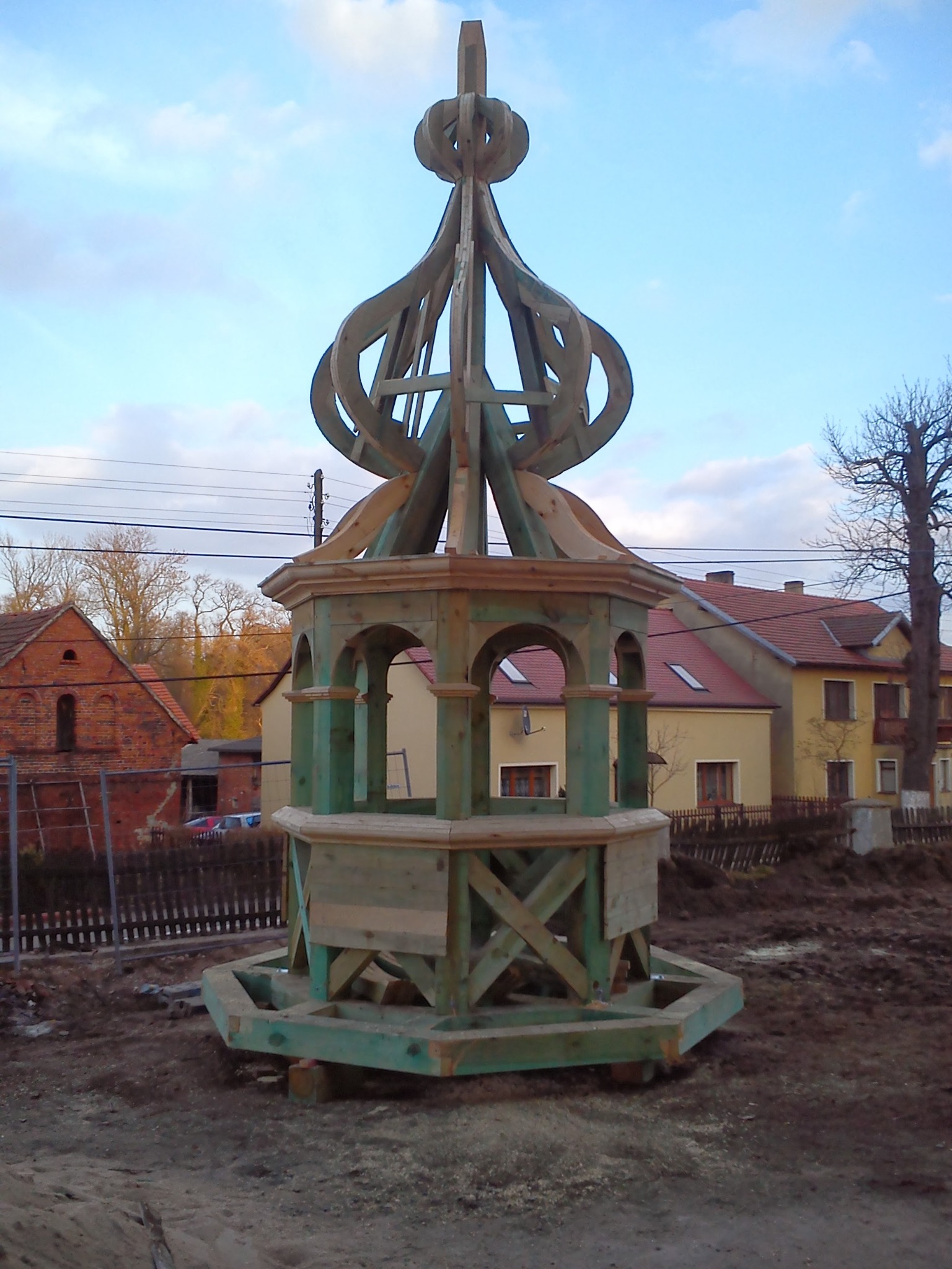 Drewniana konstrukcja altany ogrodowej w trakcie budowy, z widocznymi elementami więźby i dekoracyjnym zwieńczeniem na tle zabudowań mieszkalnych.