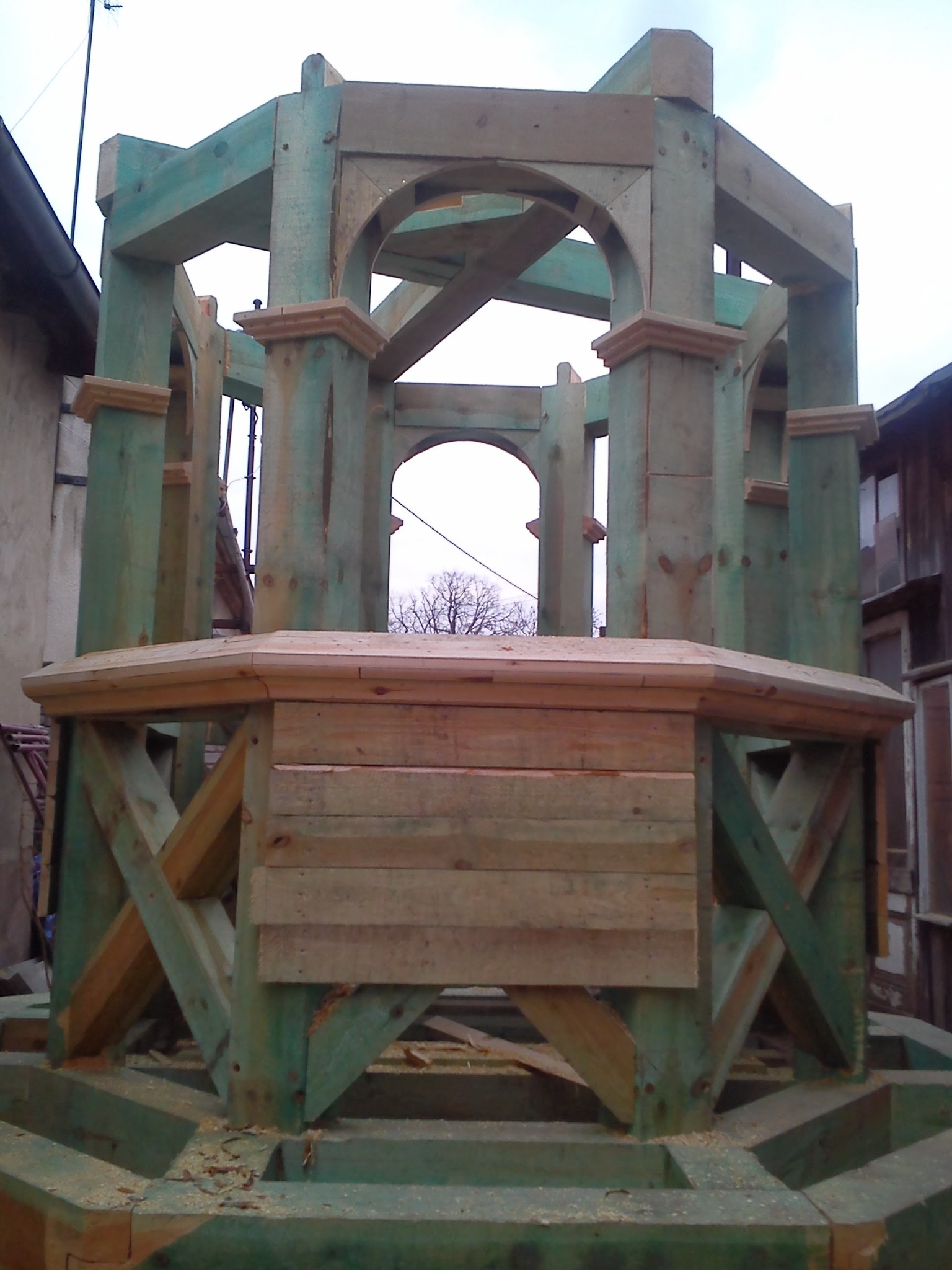 Surowa, drewniana konstrukcja altany z łukowymi przejściami, pomalowana na zielono, w trakcie budowy na tle zabudowań.